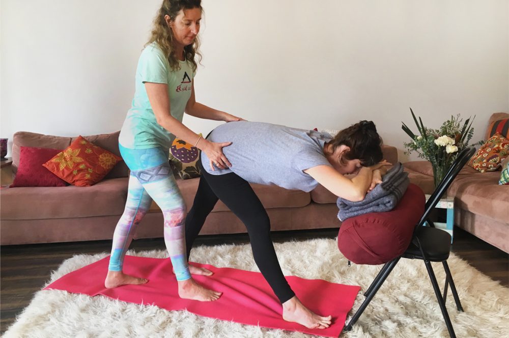Ana giving Sophie a gentle hip adjustment in a Supported Parsvottanasana with forehead resting on forearms supported by a bolster and blanket on the seat of a chair