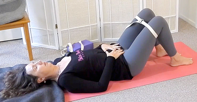 Ana Davis lying on her peach coloured yoga mat in Constructive Rest pose with strap around thighs, feed hip-width and hands resting on lower abdomen, wearing a black long sleeved top and grey leggings