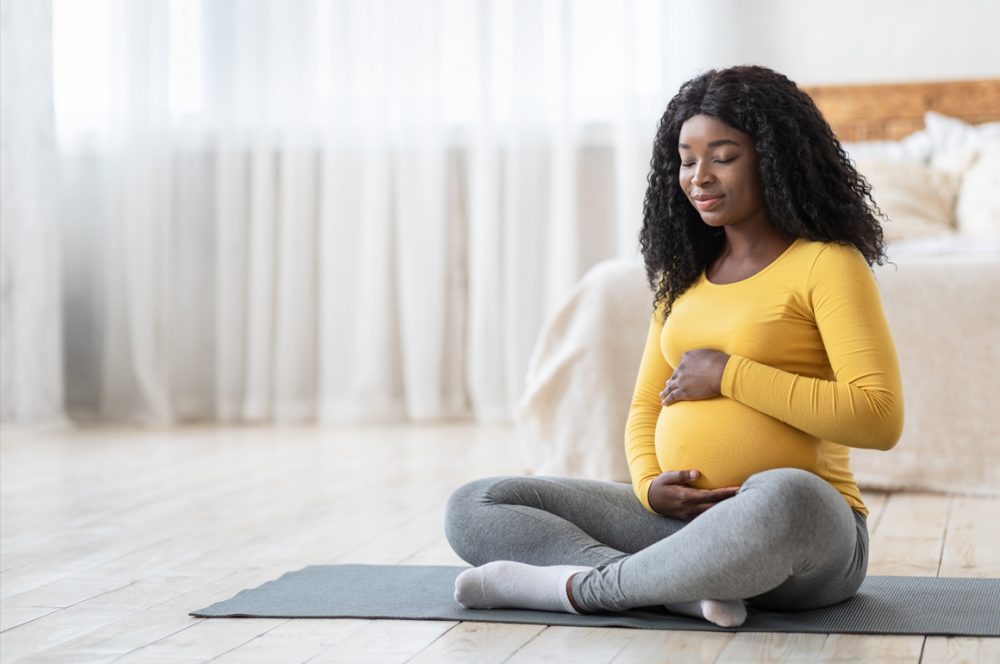 A Black woman wearing a yellow top and grey leggings sitting on a yoga mat holding her pregnant belly with eyes closed, with a bed in the background