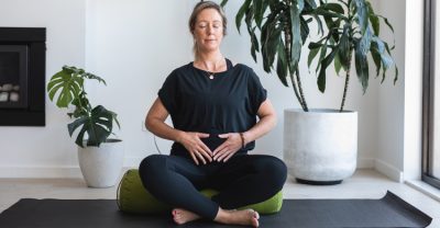 Tanya Neate, a white woman with blonde hair wearing a black top and black leggings sitting on a green bolster, legs crossed with hands on womb space. There are pot plants in the background.